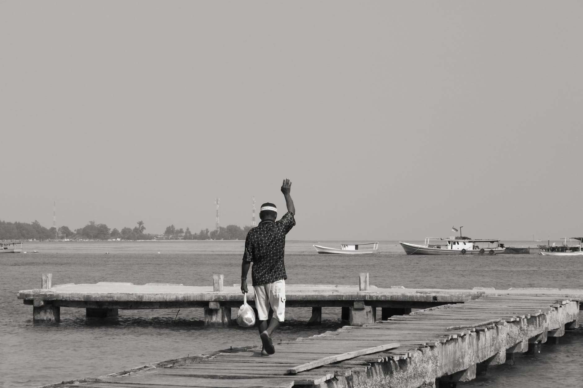 grayscale photo of a man walking on wooden dock waving