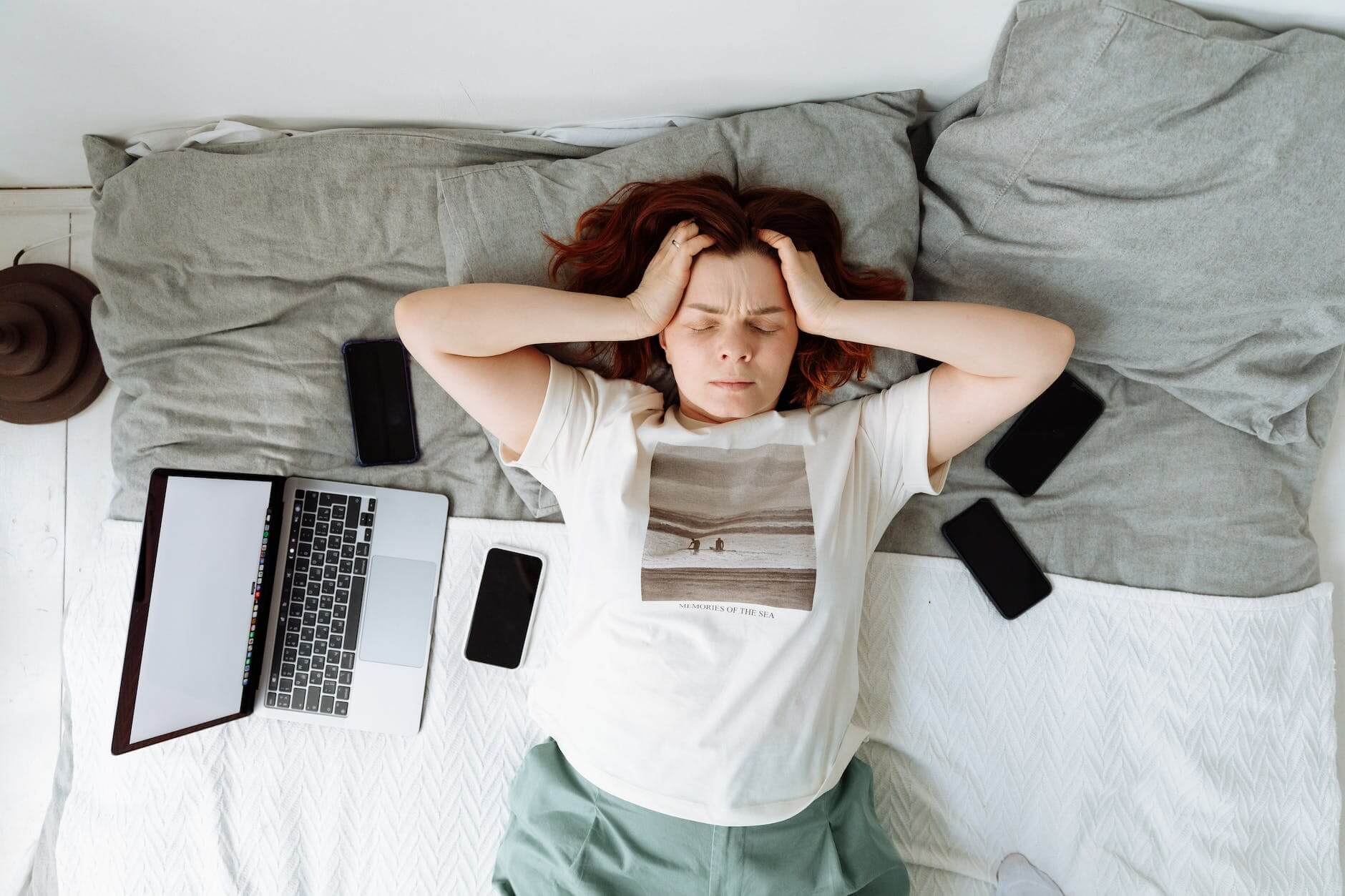 a stressed woman lying on a bed beside cellphones and a laptop
